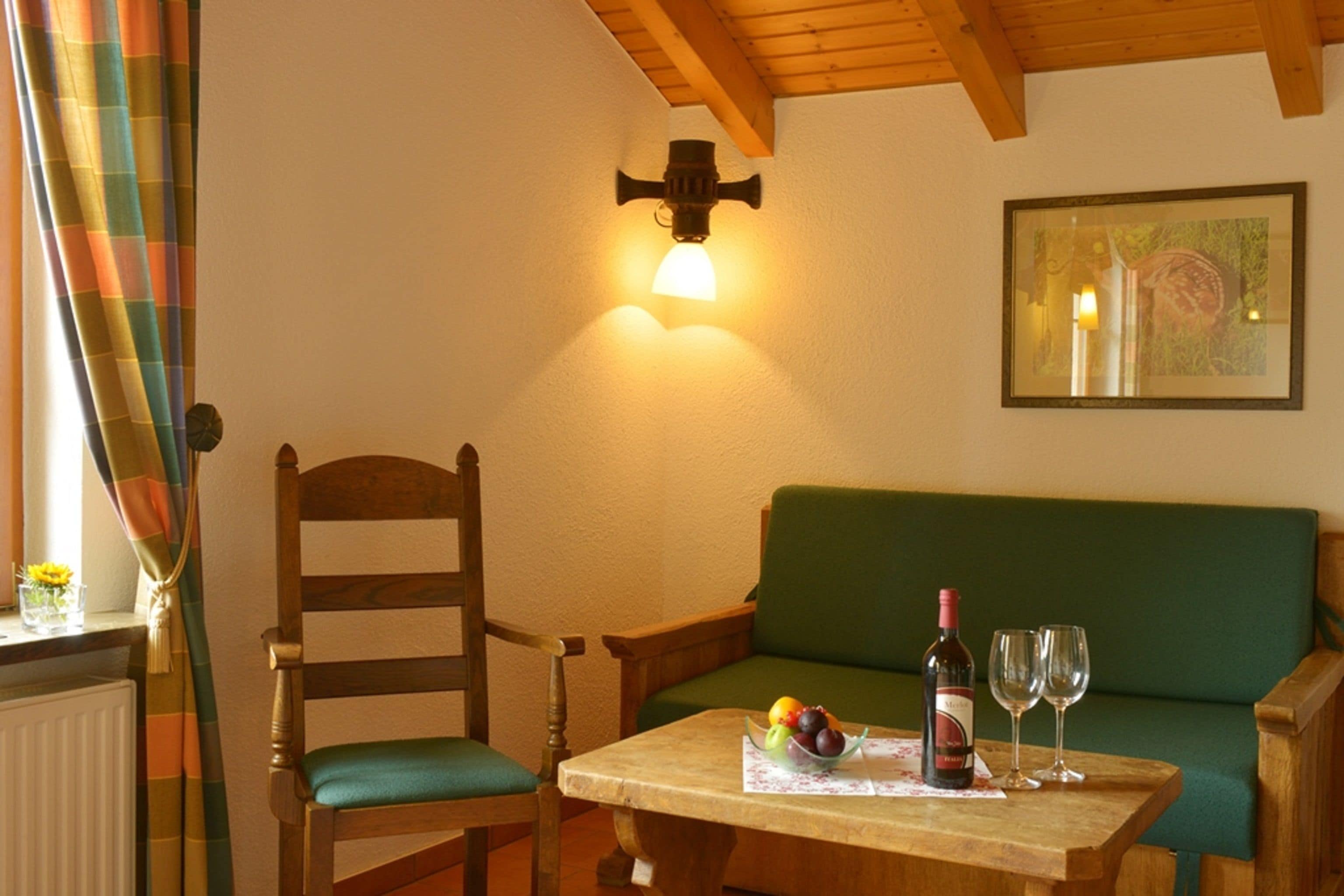 A cozy room with a wooden chair, a green sofa, a table with a bottle of wine and glasses, and a wall-mounted light fixture.