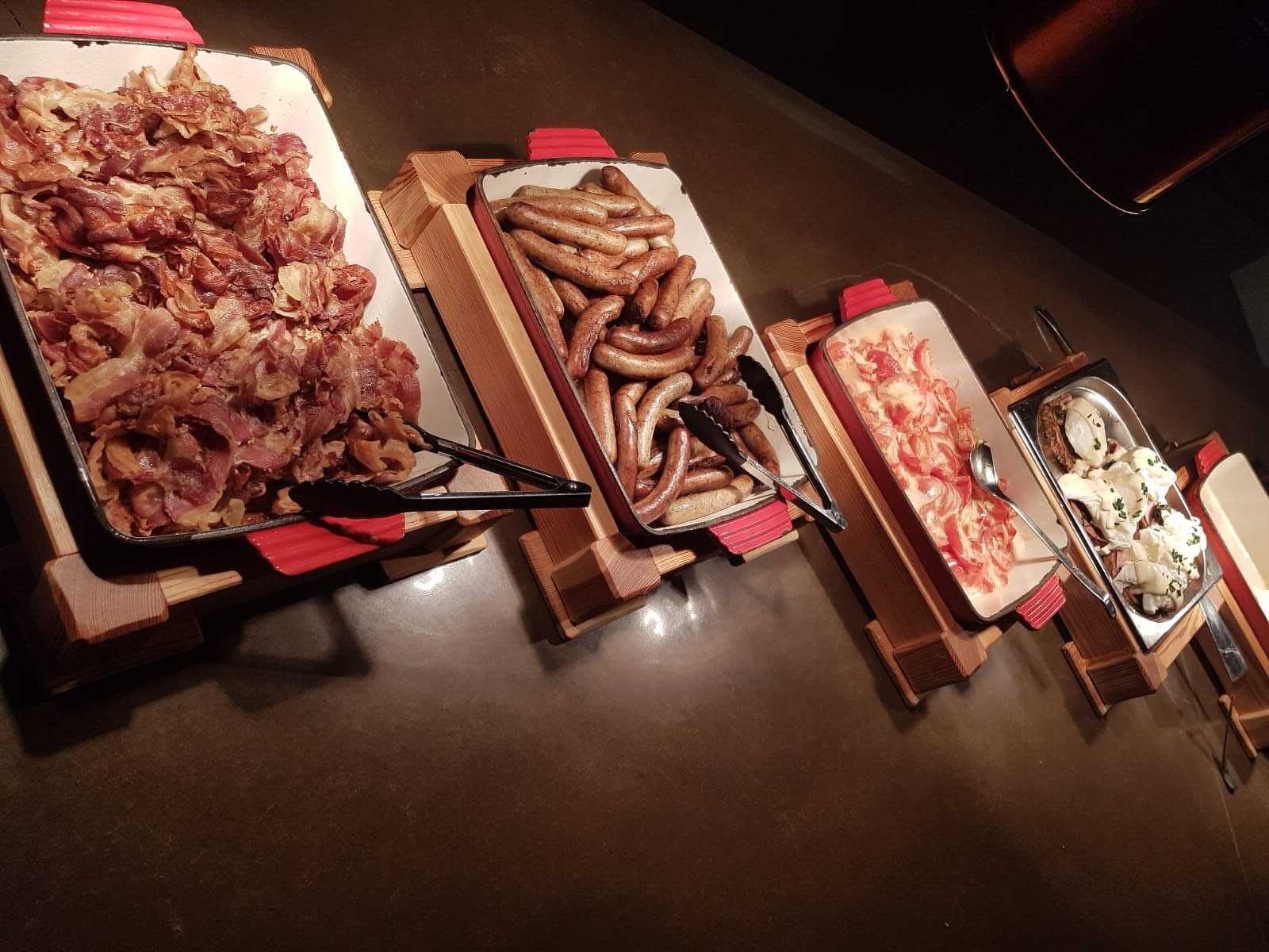 A buffet table with various food items including bacon, sausages, and a pizza.