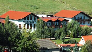 Un hotel europeo tradicional con tejados de tejas rojas, rodeado de abundante vegetación.