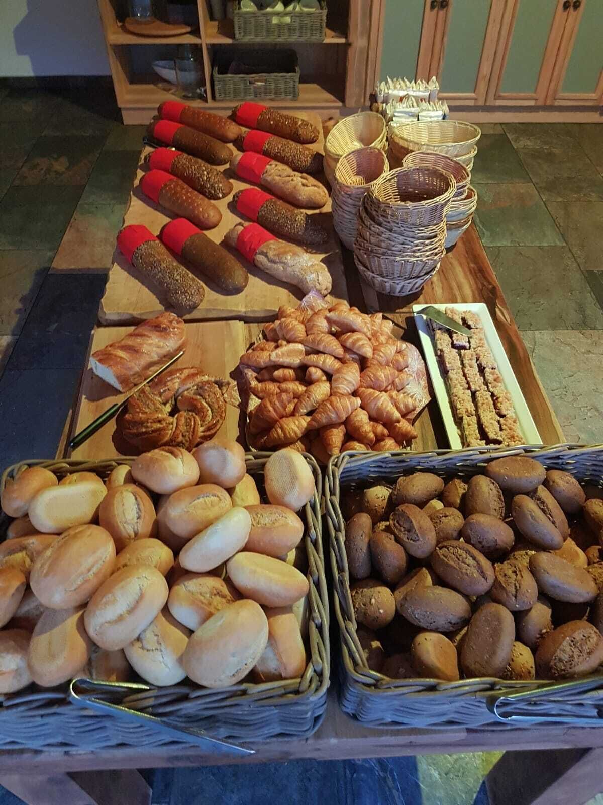 A variety of baked goods displayed on a wooden table, including breads, rolls, and pastries, with baskets and containers for serving.