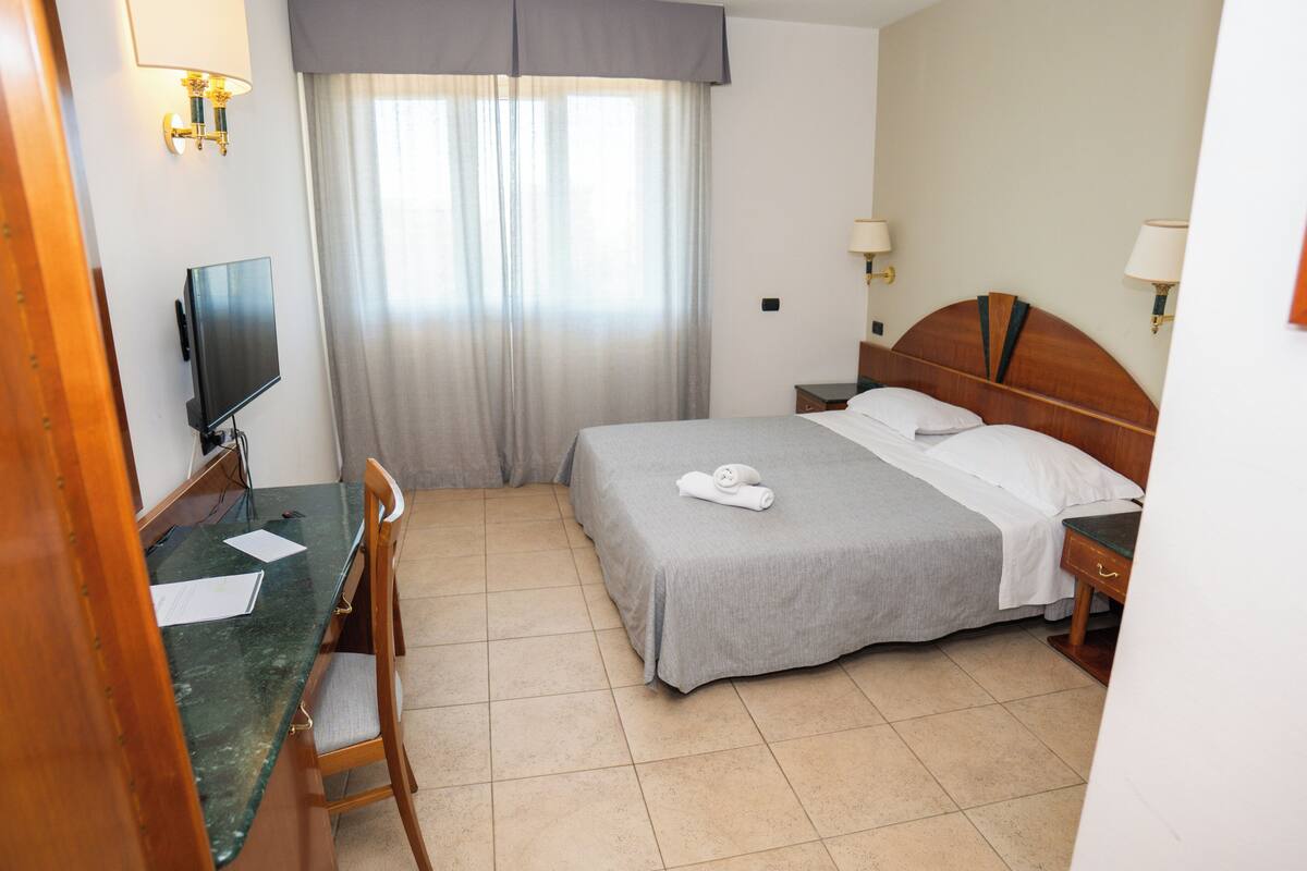 Ein Hotelzimmer mit einem Bett, einem Schreibtisch mit Papieren, einem Fernseher und einem Fenster mit Vorhängen.