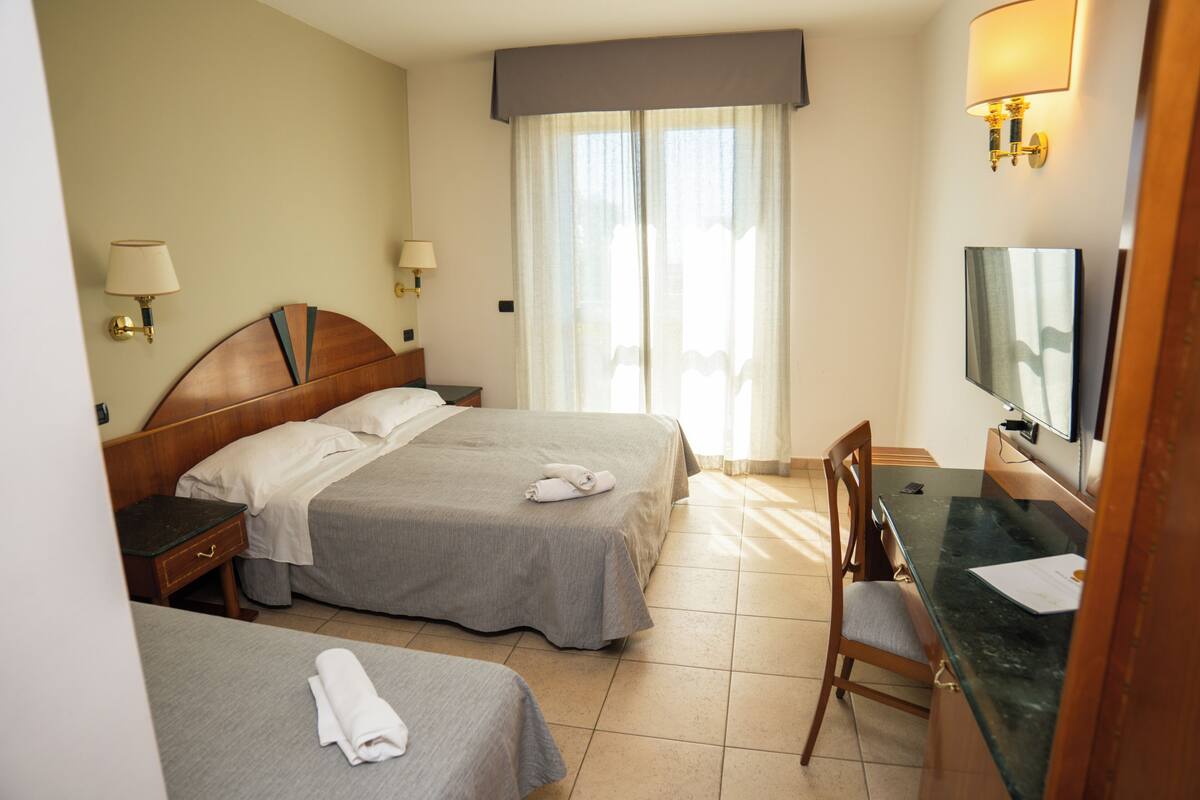 Ein Hotelzimmer mit zwei Betten, einem Schreibtisch, einem Stuhl, einem Fernseher und einem Fenster mit Vorhängen.