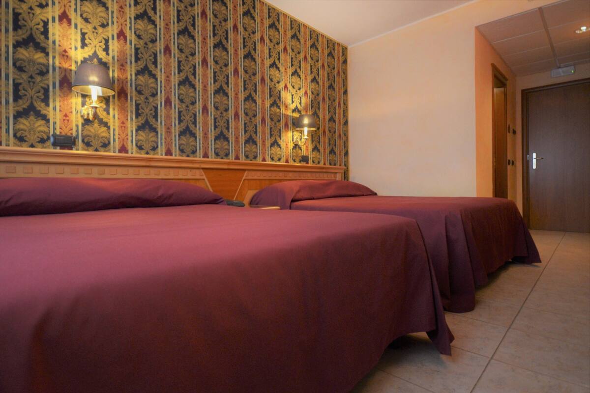 Ein Hotelzimmer mit drei Betten, jeweils mit einer roten Bettdecke, und zwei Wandlampen.