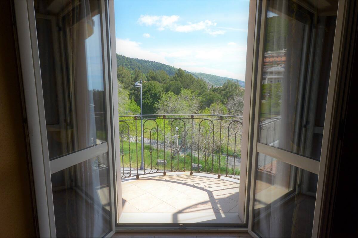 Ein Balkon mit Metallgeländer, Fliesenboden und Blick auf eine grüne Landschaft mit Bäumen und Hügeln.