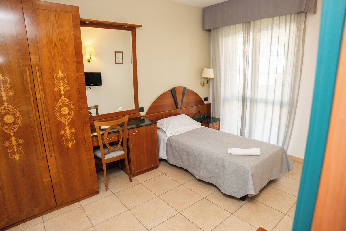 Ein Hotelzimmer mit Bett, Schreibtisch, Stuhl, Kleiderschrank, Spiegel, Fernseher und einem Fenster mit Vorhängen.