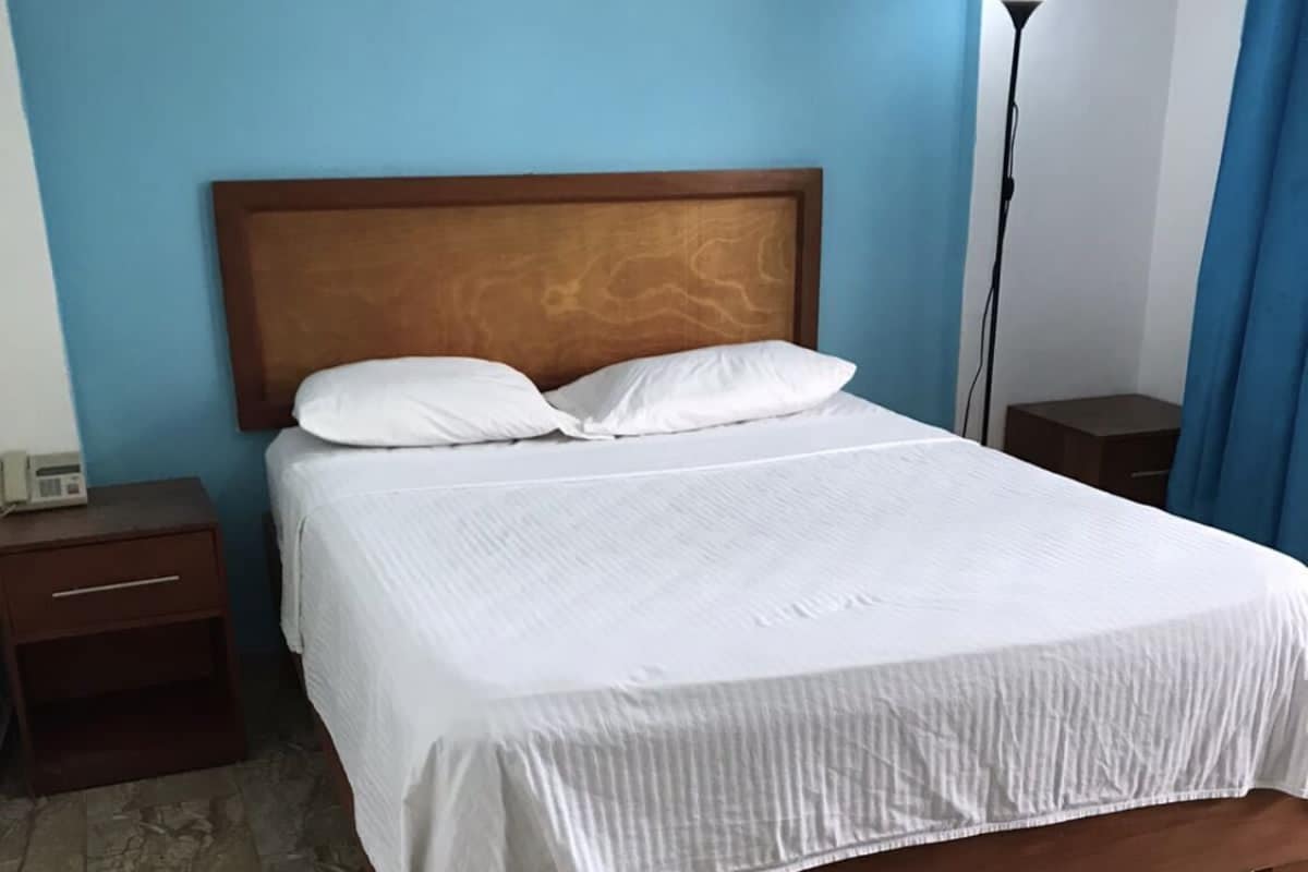 Een bed met een houten hoofdbord, witte beddengoed, een nachtkastje, een lamp en een blauw gordijn.