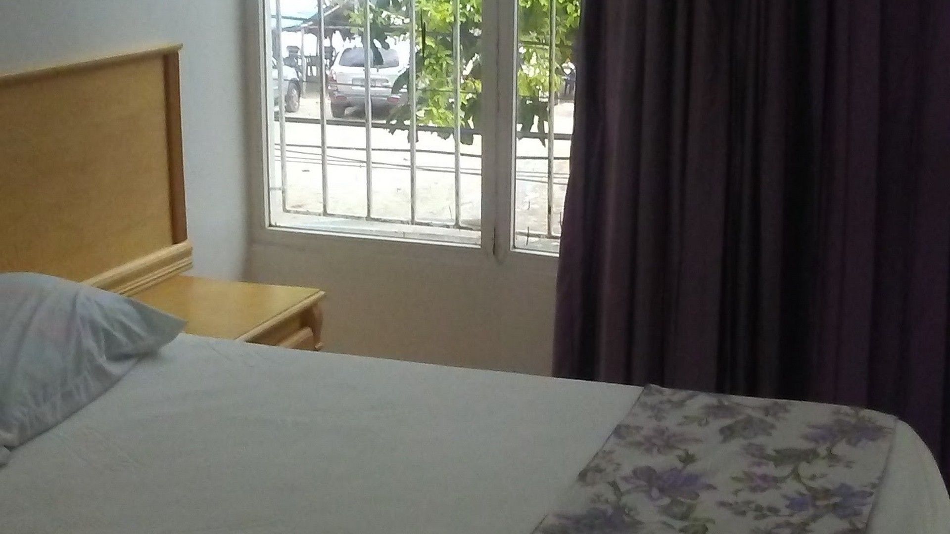 A bedroom with a bed, a nightstand, a window with bars, and a view of the outside.