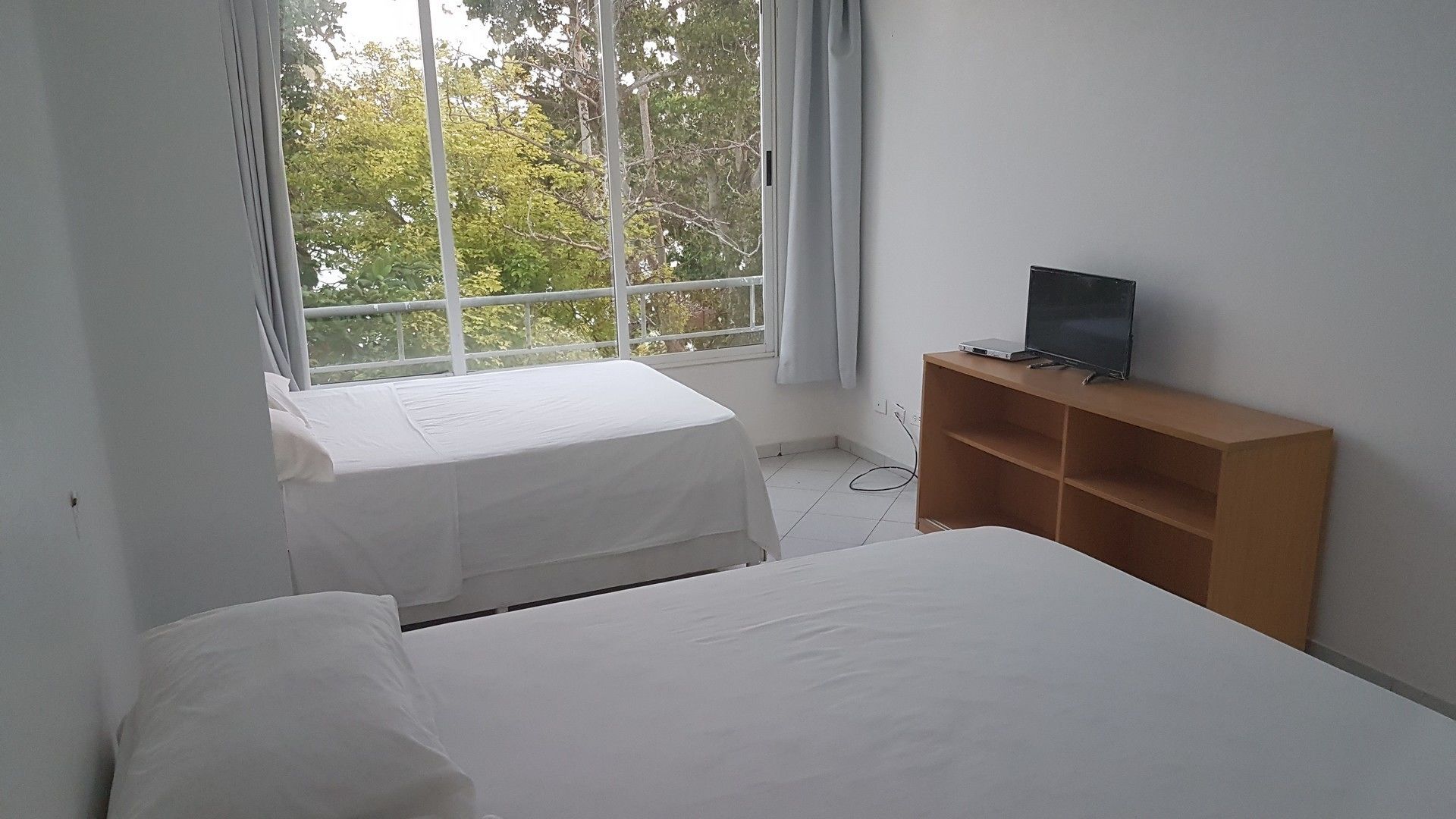 A bedroom with two beds, a TV on a wooden shelf, and a window with curtains.