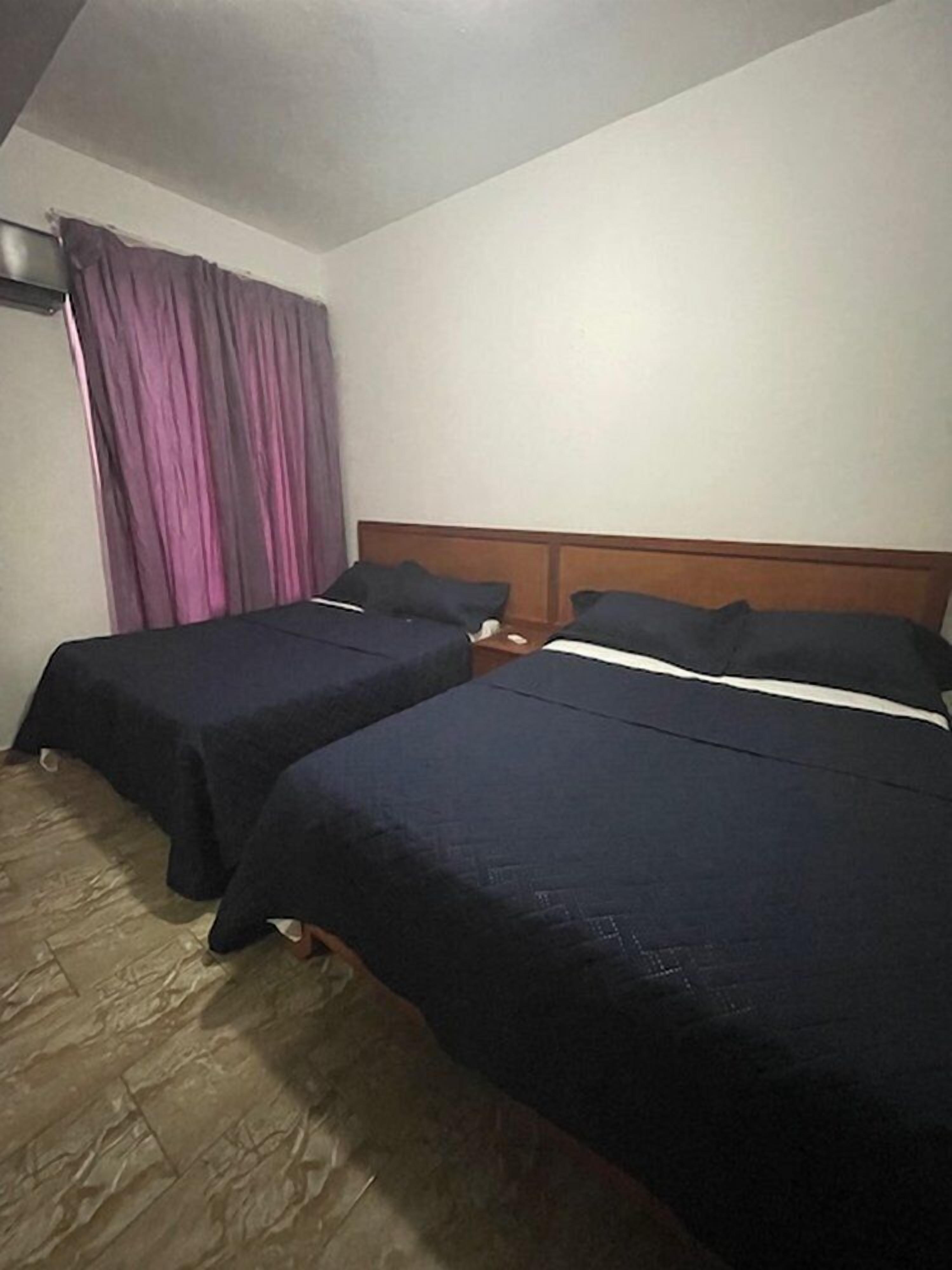 Habitación de hotel con dos camas, cabecera de madera, mesita de noche y ventana con cortinas moradas.