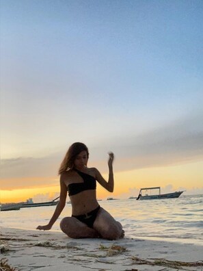 Una persona sentada en la playa al atardecer, con un barco al fondo.