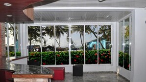 Eine moderne Lobby mit Steinboden, einem hölzernen Empfangsbereich und großen Fenstern, die einen Blick auf eine angelegte Grünfläche mit Palmen und ein Gebäude im Hintergrund bieten.