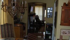 A dining area with a wooden table, chairs, and a chandelier. There is a kitchen area in the background with a person visible. - Turner Falls (Davis)