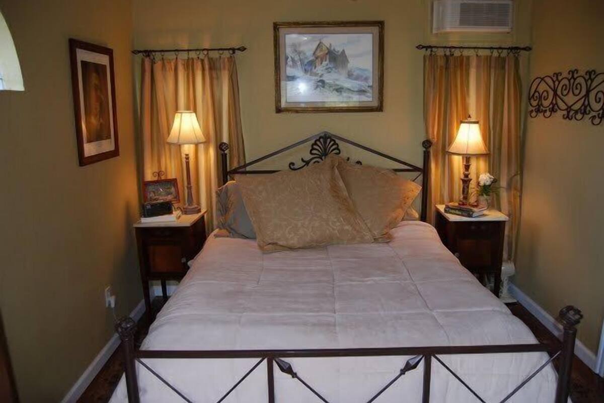 A bedroom with a bed, two bedside tables, a nightstand, a painting on the wall, and curtains.