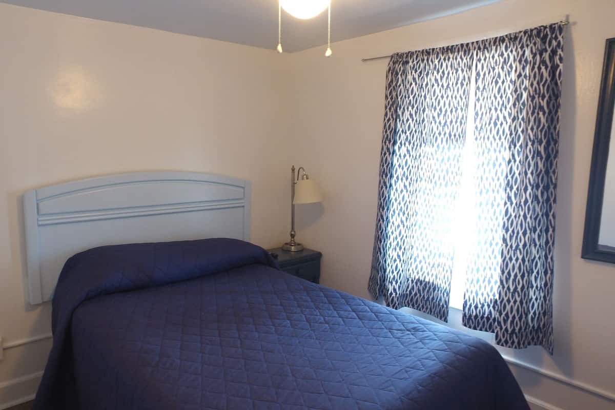 A bedroom with a bed, a bedside table with a lamp, and a window with patterned curtains.
