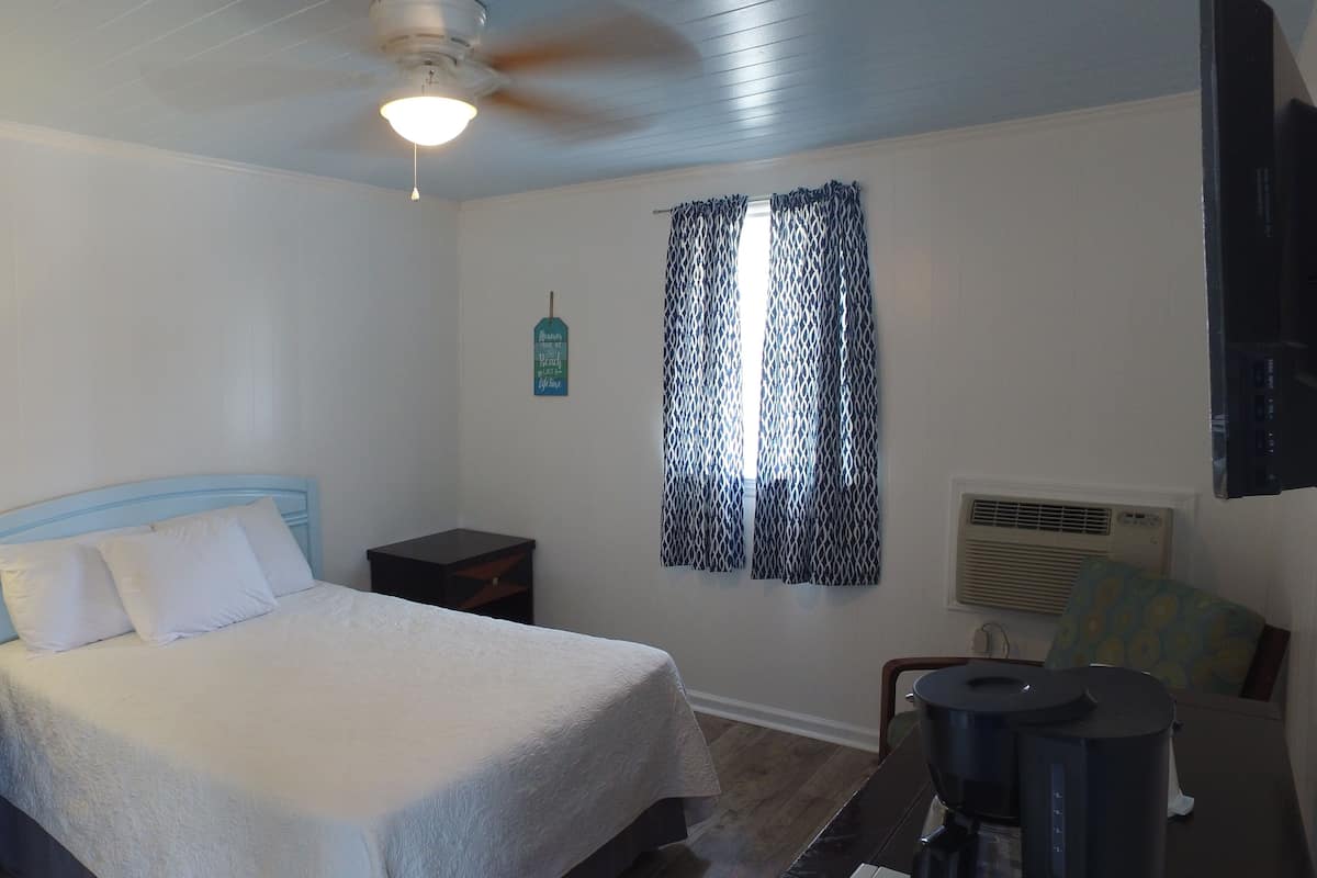 A bedroom with a bed, nightstand, lamp, ceiling fan, window with patterned curtains, air conditioning unit, and a coffee maker.