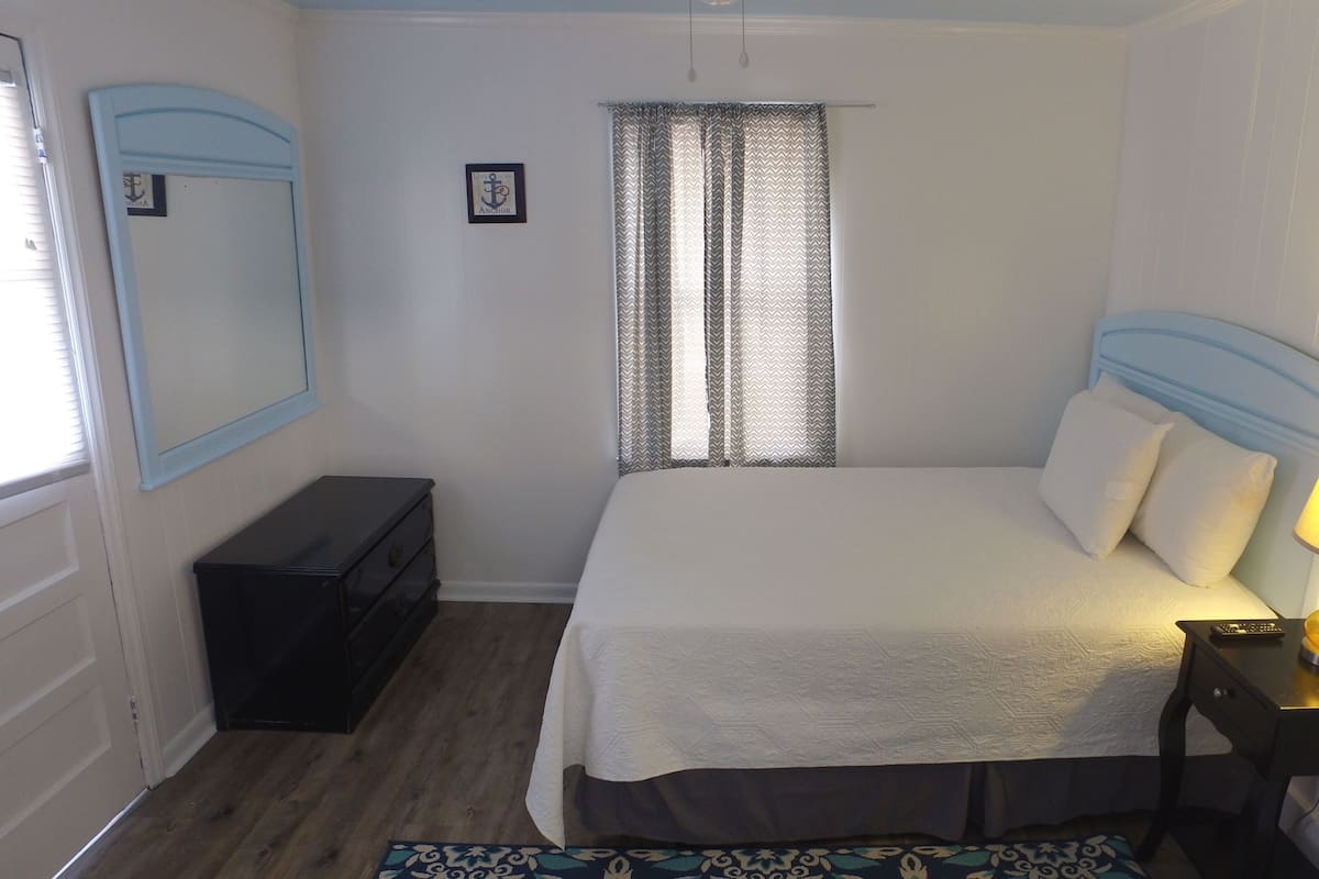 A bedroom with a bed, a nightstand with a lamp, a dresser, a window with curtains, and a framed picture on the wall.