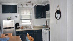 A compact kitchen with dark cabinets, a white refrigerator, a stove, and a microwave. There is a dining table with chairs and a window with blinds. - Midtown Cottages (Myrtle Beach)