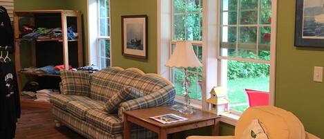 A living room with a plaid sofa, a yellow armchair, a wooden table, and a window with a view of greenery.