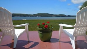 Two white chairs facing a scenic view of a lake with a green field and a pot of red flowers in the center. - The Beach Inn Motel (Munising)
