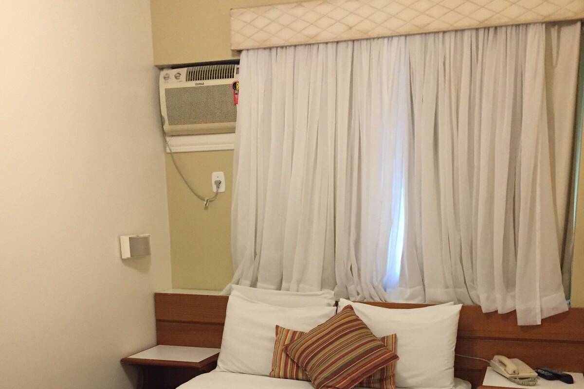 A hotel room with a queen-sized bed, a bedside table with a phone and remote, a wall-mounted air conditioner, and a window with white curtains.