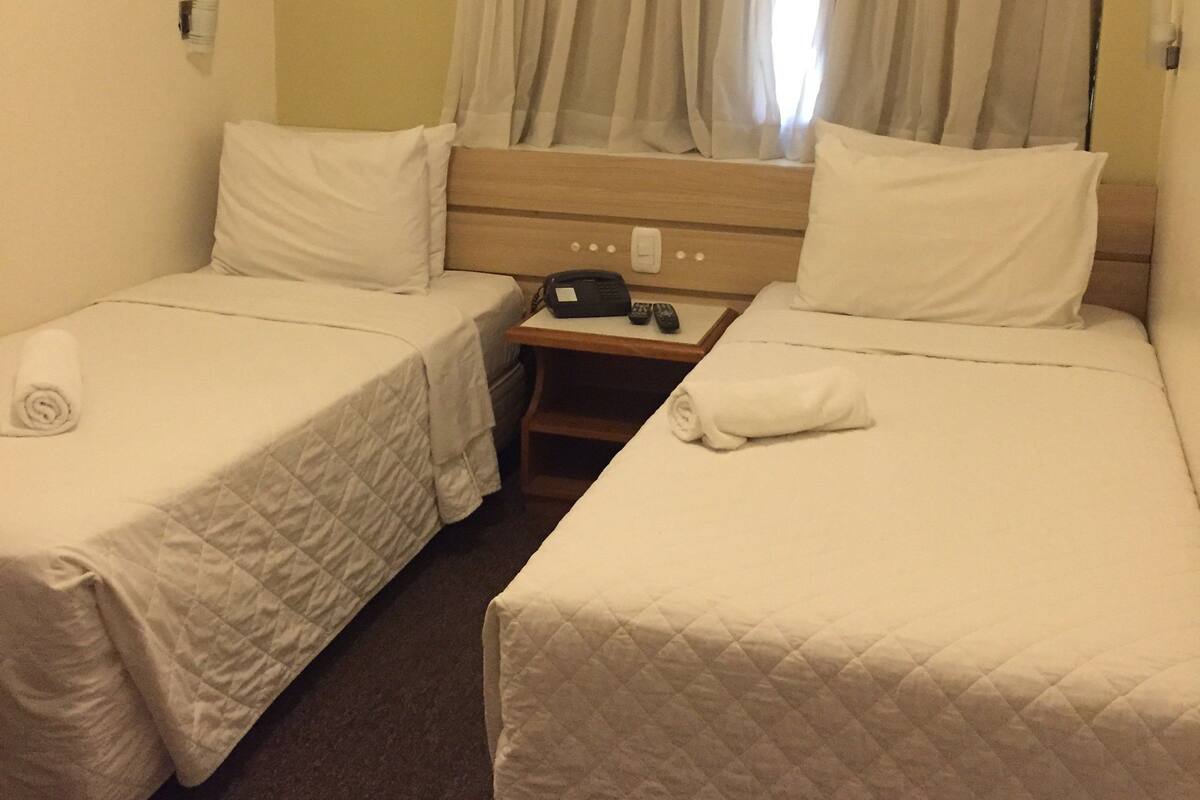 A hotel room with two beds, a bedside table with a phone and remote, and an air conditioning unit above the window.
