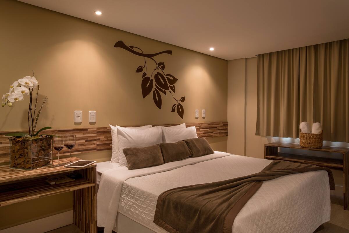 A modern hotel room with a large bed, a wooden headboard, a wall-mounted plant, and a bedside table with a vase and wine glasses.