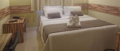 A hotel room with a bed, a wooden side table, a telephone, and a wall-mounted air conditioner.