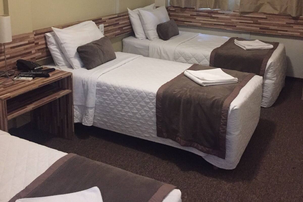 A hotel room with two beds, a wooden headboard, a bedside table with a lamp, and a window with curtains.