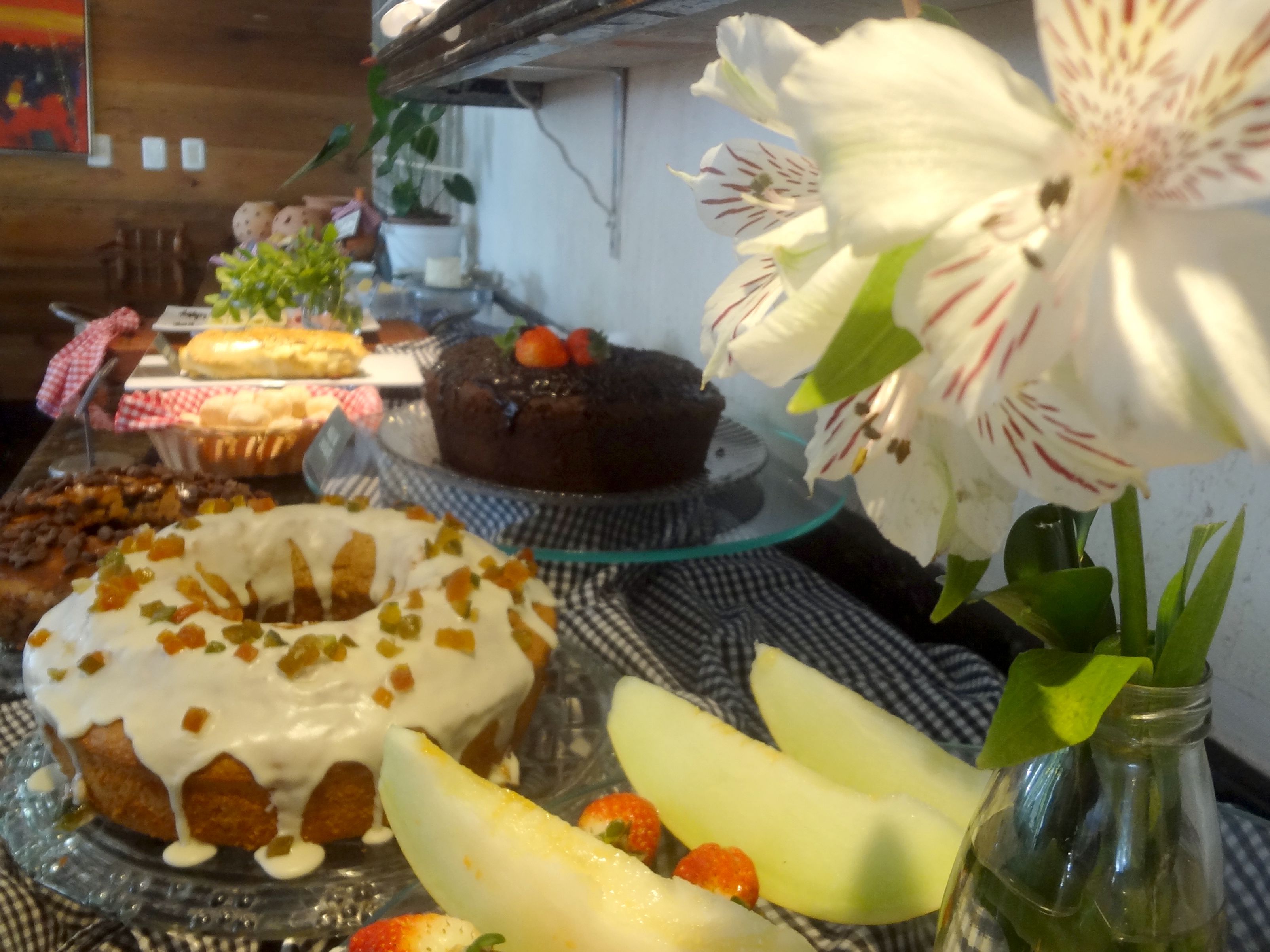 A table with various desserts, including a cake with strawberries, a donut with white icing, and a chocolate cake with a strawberry on top. There is also a vase with white flowers and a glass bottle.