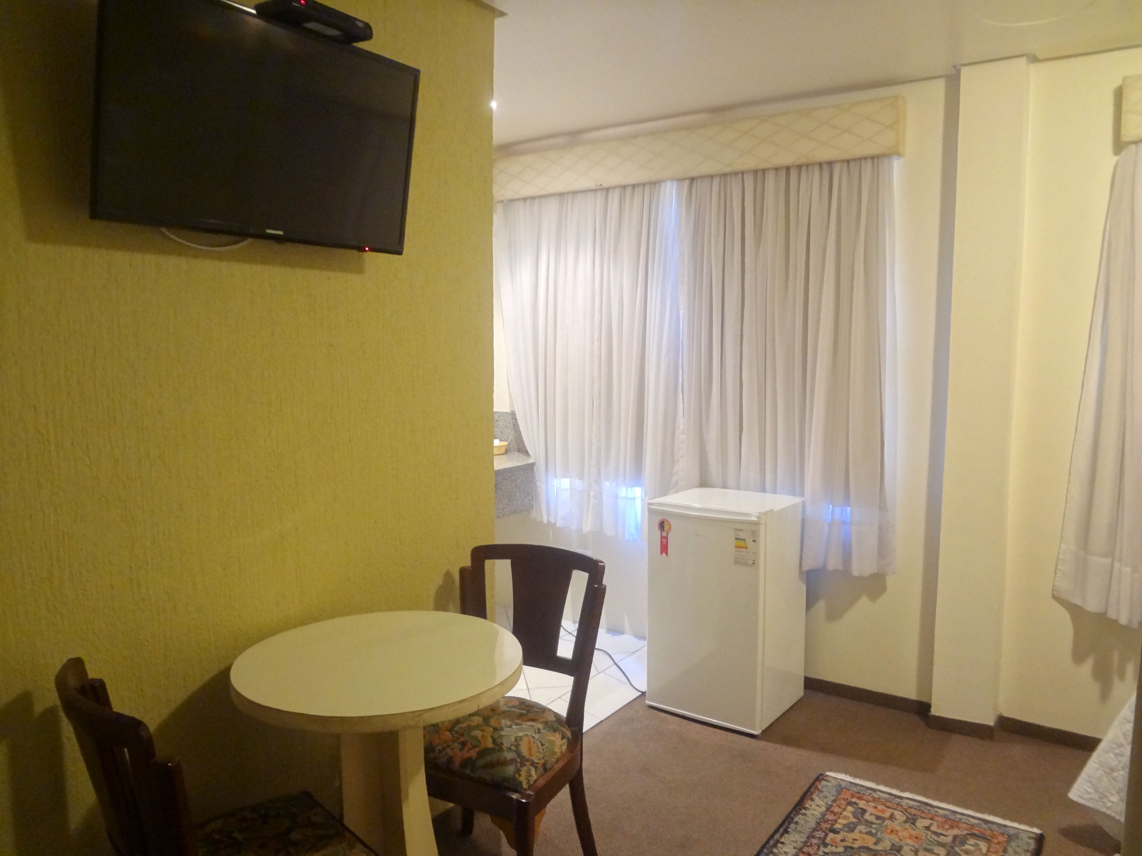 Ein kompaktes Hotelzimmer mit einem runden Tisch, einem Stuhl, einem an der Wand montierten Fernseher, einer kleinen Kühlbox und einem Fenster mit Vorhängen.