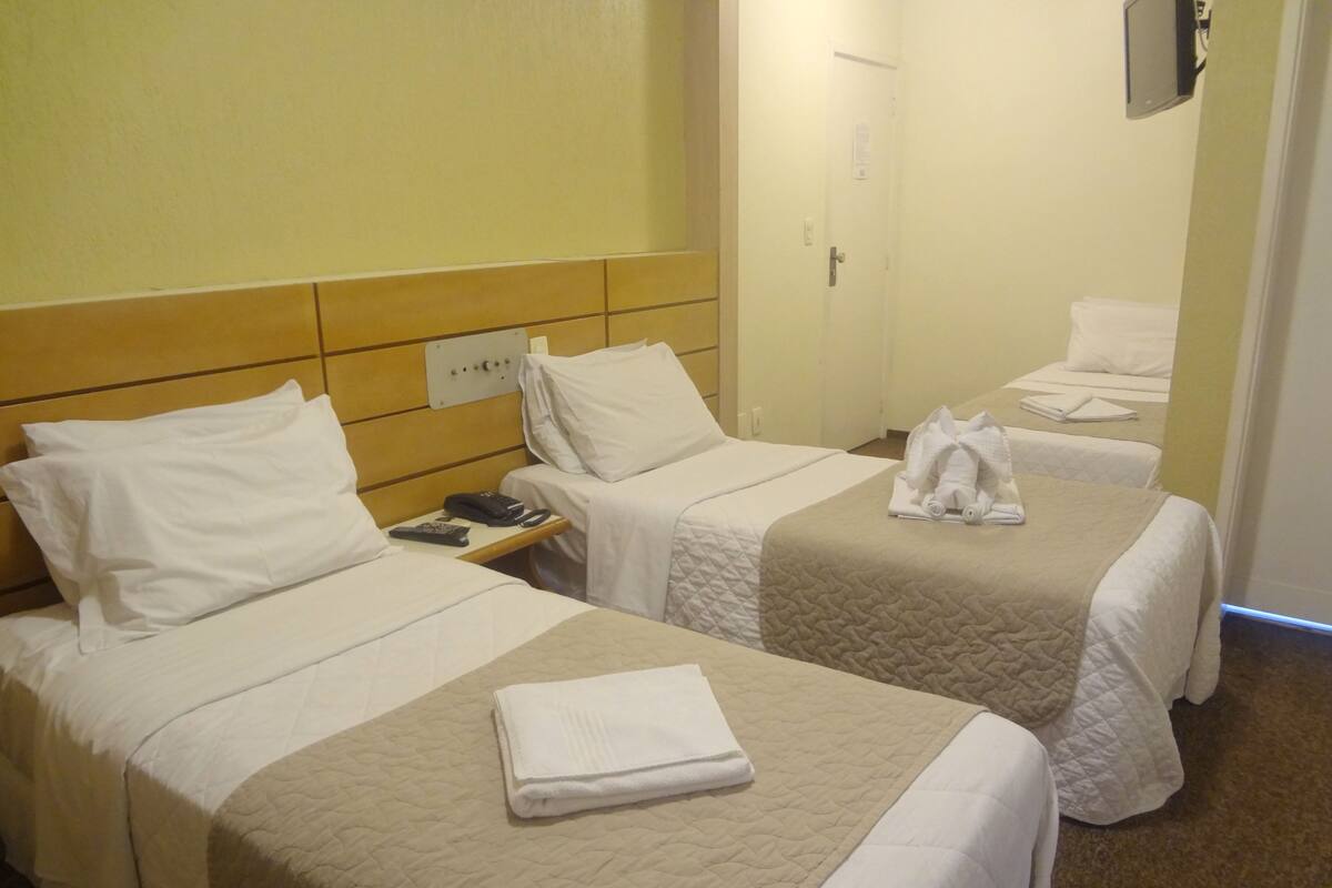 A hotel room with two single beds, each with a white bedspread and pillows. There is a folded towel on the bed in the foreground. A nightstand with a phone and remote control is visible.