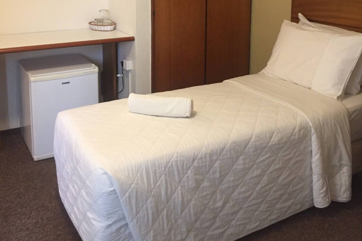 A hotel room with a bed, a wooden wardrobe, a small refrigerator, a mirror, and a bedside table with a basket.