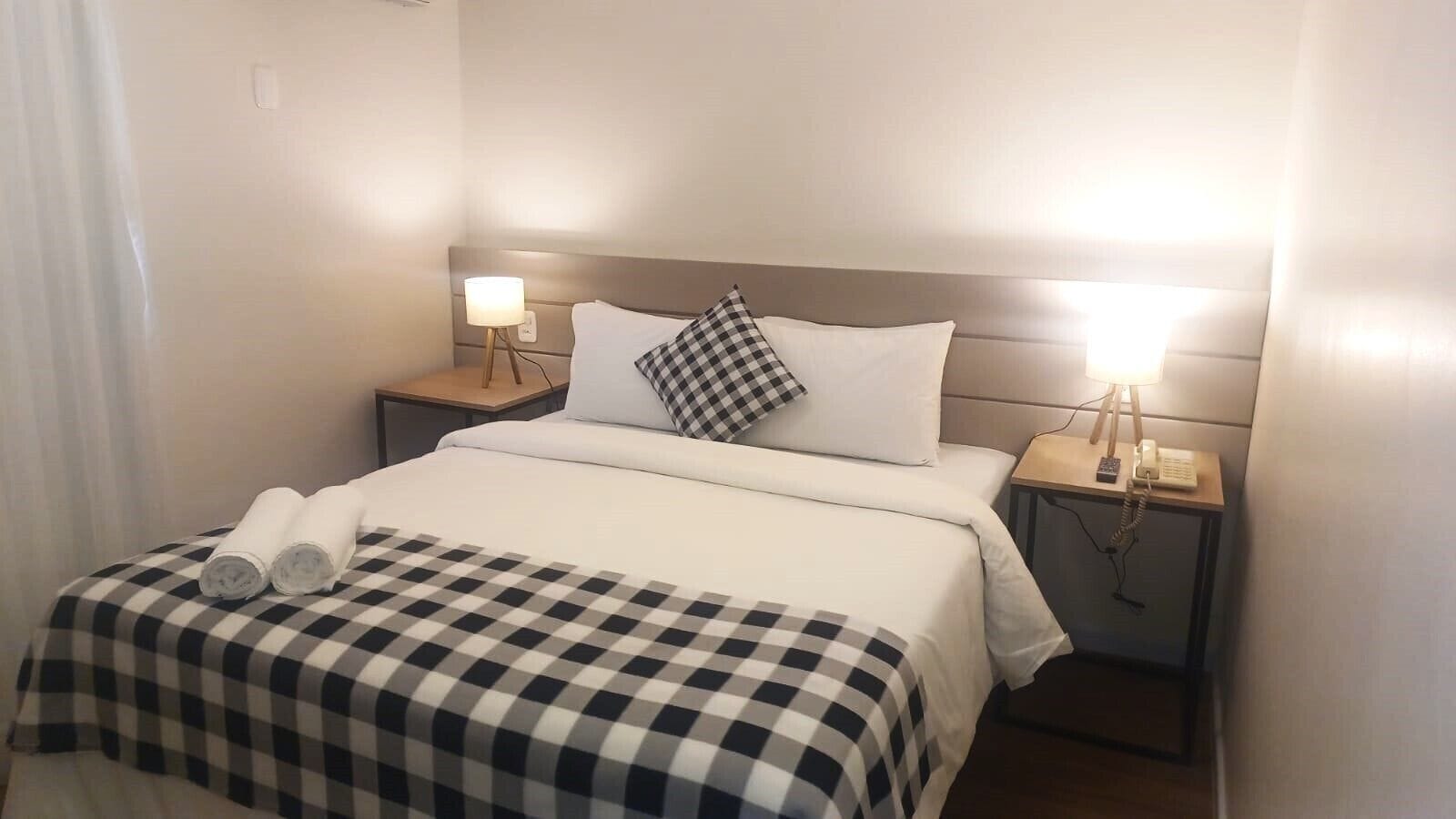 Habitación de hotel con una cama, dos mesitas de noche con lámparas, una colcha a cuadros y una toalla enrollada sobre la cama.