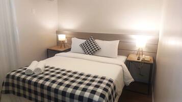 Quarto de hotel com uma cama, duas mesas de cabeceira com luminárias, um edredom xadrez e uma toalha enrolada sobre a cama.