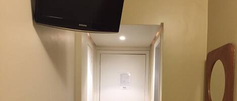 Un pasillo estrecho con una puerta blanca al final, un refrigerador pequeño, un espejo y un televisor montado.