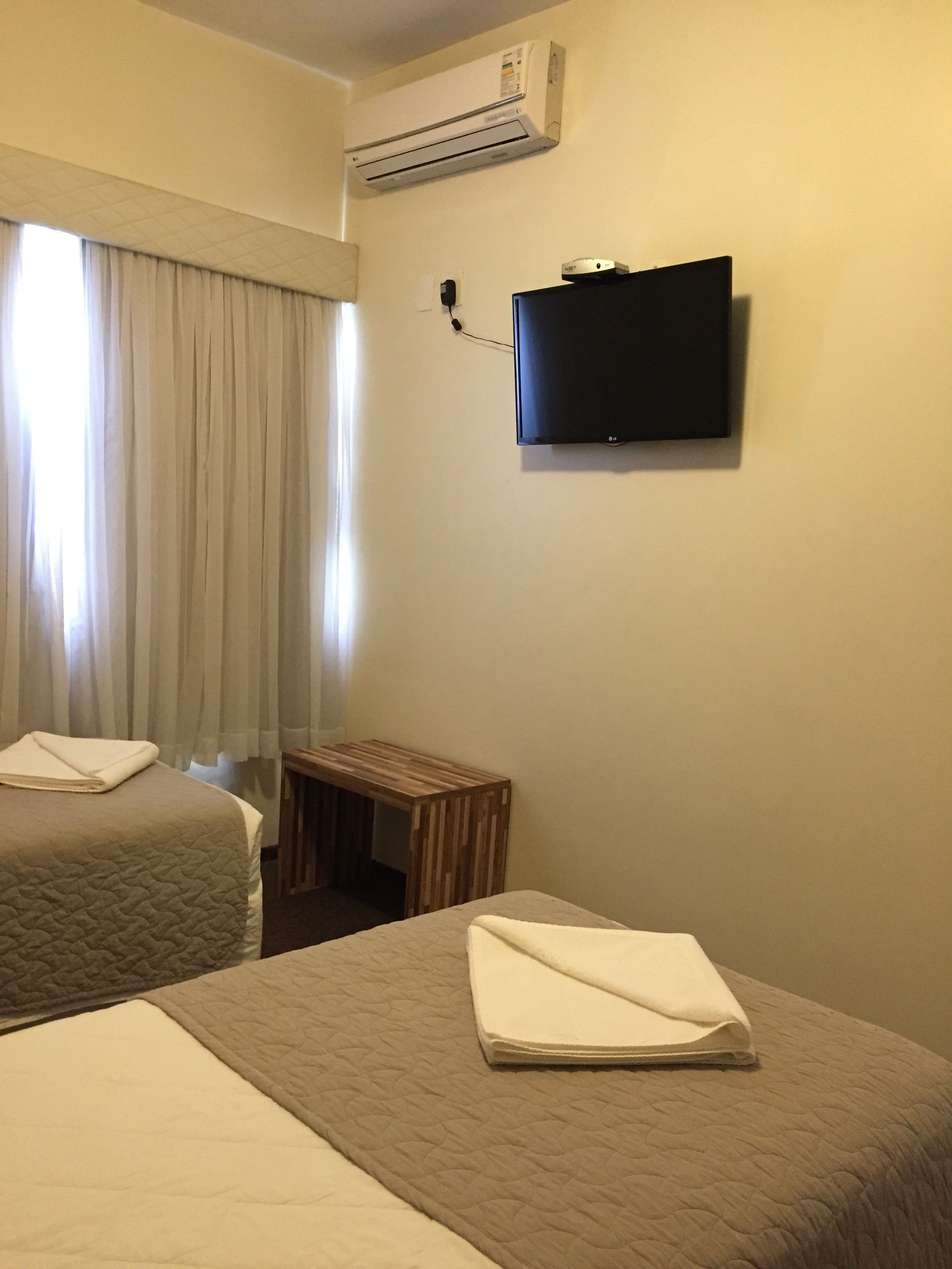 A hotel room with two beds, a TV mounted on the wall, an air conditioner, and a small wooden table.