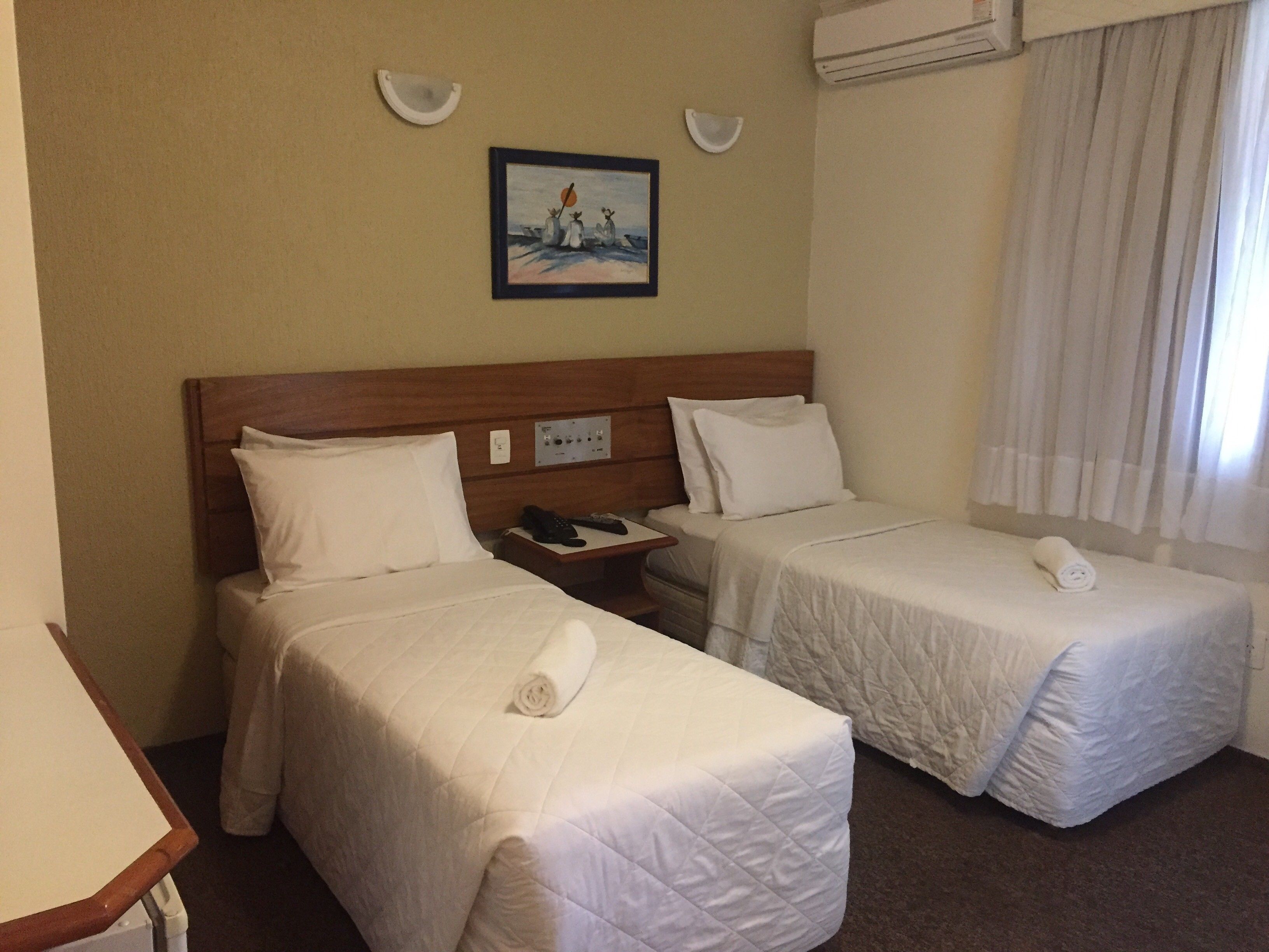 Ein Hotelzimmer mit zwei Einzelbetten, einem Holzkopfteil, einem Bild an der Wand und einer Klimaanlage.