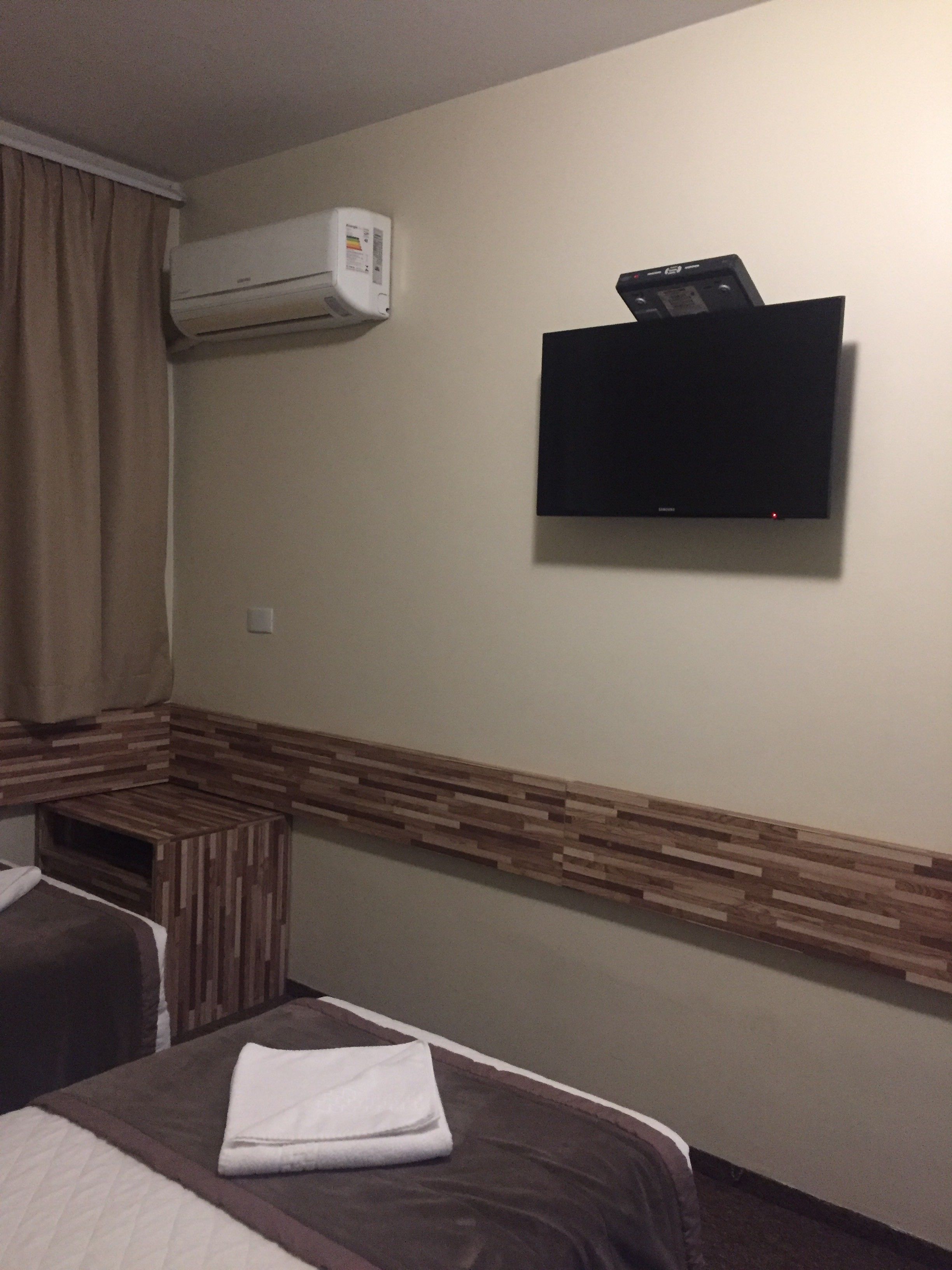 Et hotelværelse med en seng, en lille træbænk, en aircondition og et fladskærms-tv monteret på væggen.