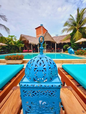 A poolside area with a decorative blue lantern, wooden lounge chairs, and thatched-roof structures in the background. - Pousada Castelinho (Aracati)