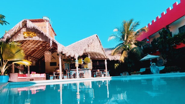 Zona de piscina en un resort con un edificio de techo de paja, palmeras y una estructura roja al fondo.