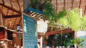 Zona de bar al aire libre de estilo rústico con un menú en pizarra que enumera bebidas y precios, una cesta de mimbre con flores y una estructura cubierta de paja.