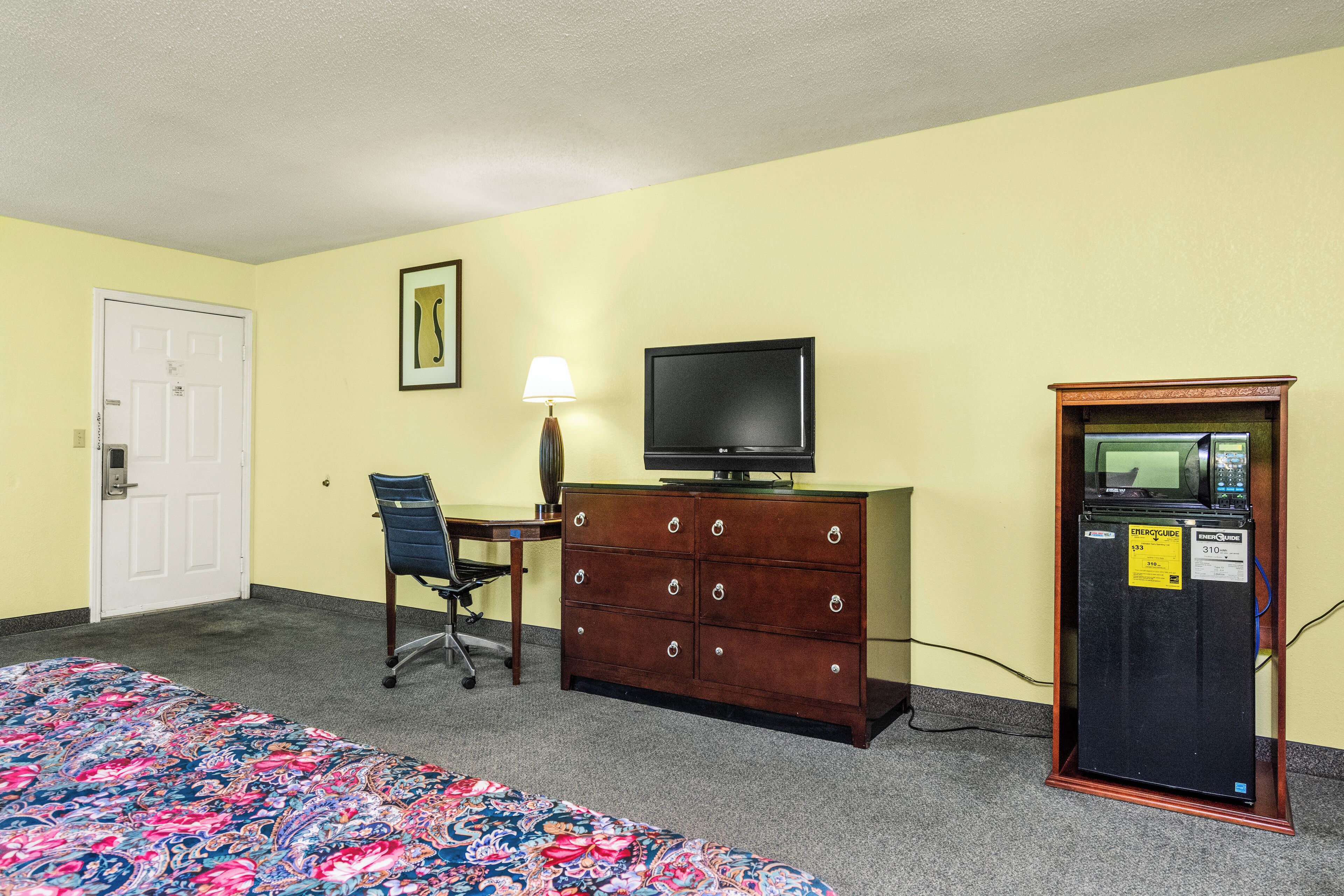A hotel room with a bed, a desk with a chair, a television, a dresser, and a refrigerator.