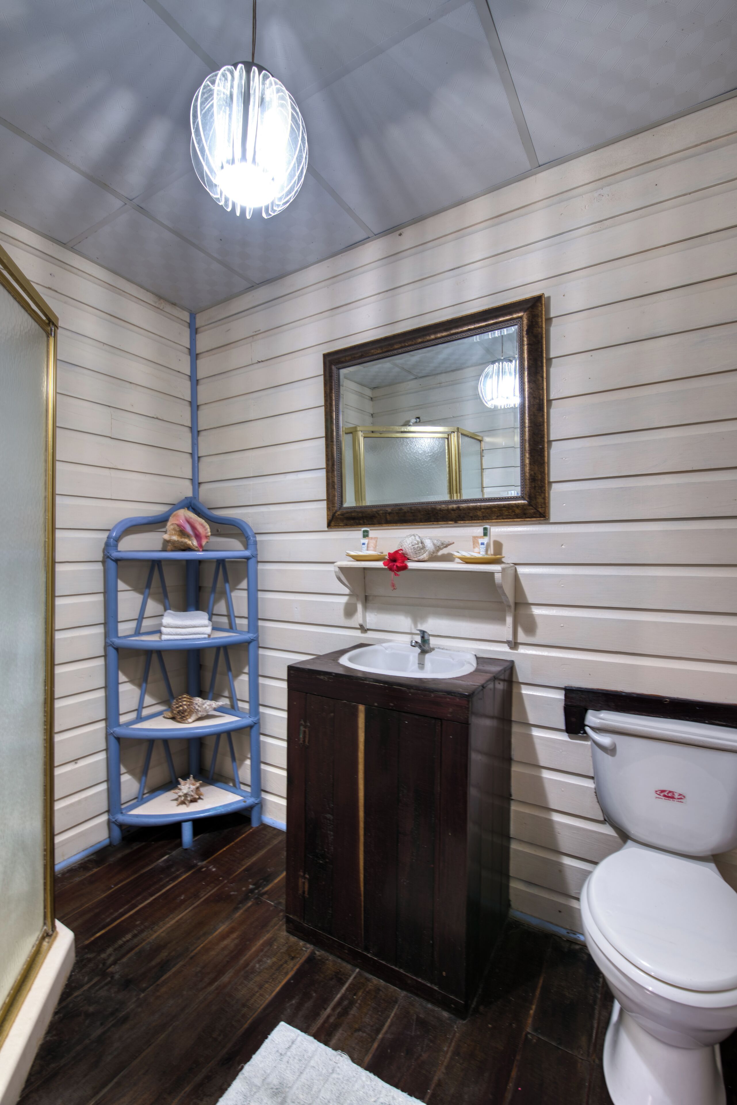 Un baño con piso de madera, un lavamanos blanco, un mueble de tocador de madera oscura, un inodoro blanco, un estante azul y un espejo con marco dorado.
