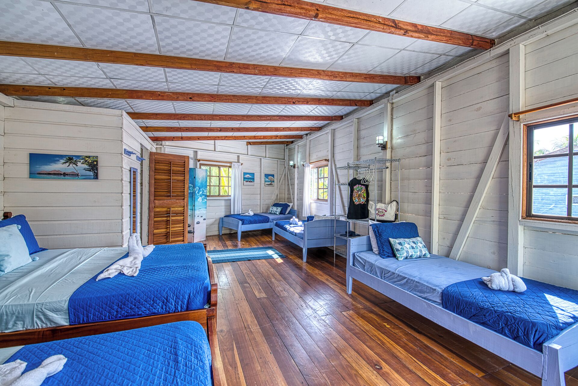 A room with wooden floors, white walls, and wooden beams on the ceiling. There are three beds with blue bedding, a metal rack with clothes, and a window with curtains.
