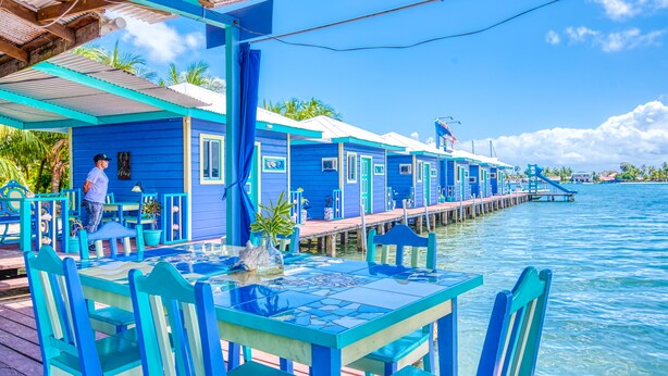 A row of blue floating houses with a dock extending into the water, a table with chairs, and a person standing by the house.