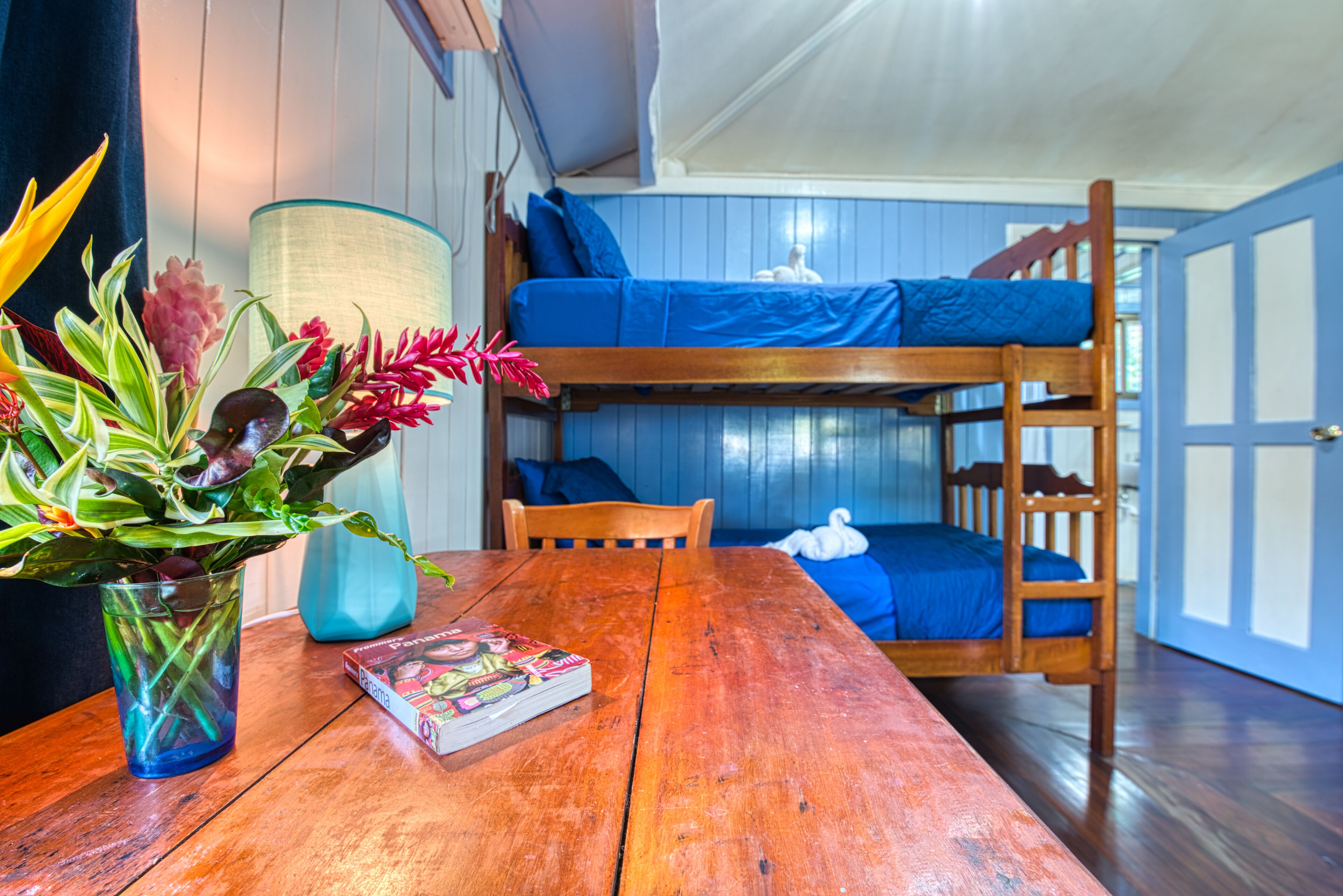 Una cama litera con ropa de cama azul, una mesa de madera con un libro y un florero, y una lámpara.