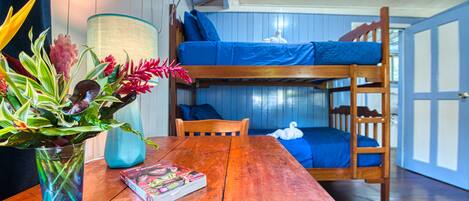 Una cama litera con ropa de cama azul, una mesa de madera con un libro y un florero, y una lámpara.