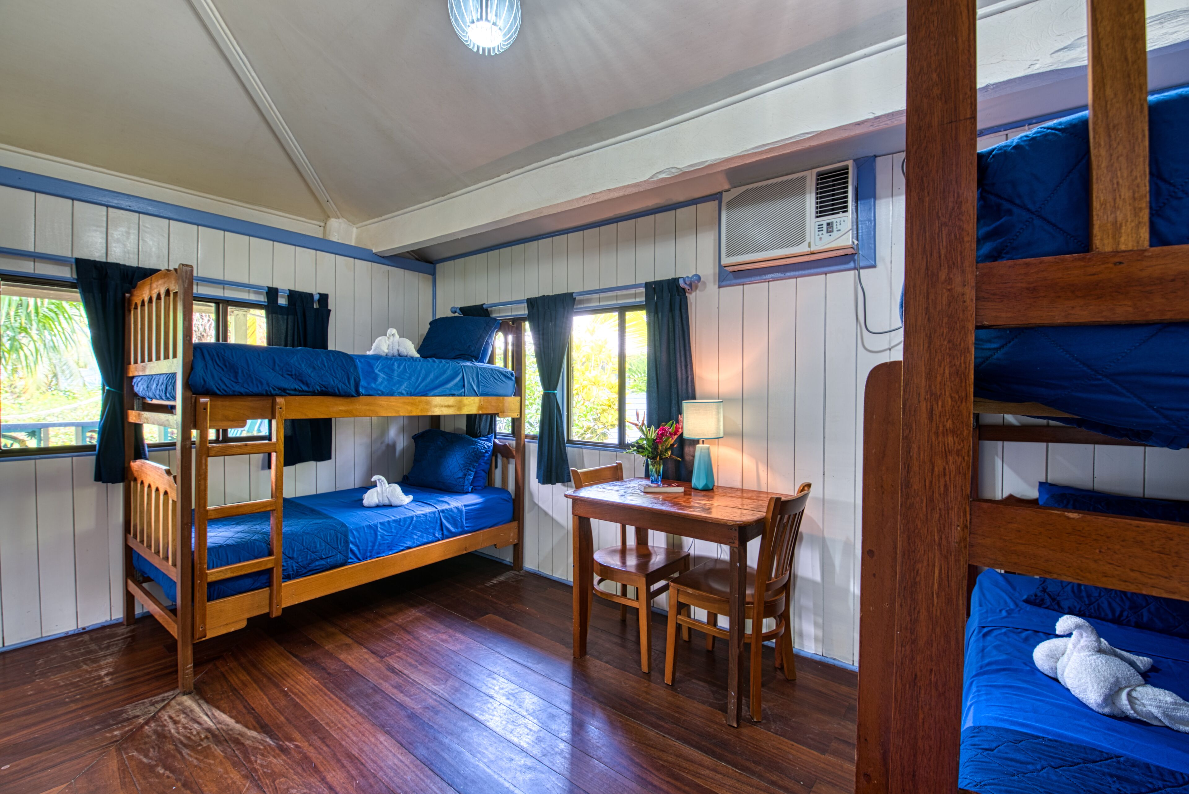 Habitación con literas, una mesa de madera y sillas, una ventana con cortinas y una lámpara de mesa pequeña.