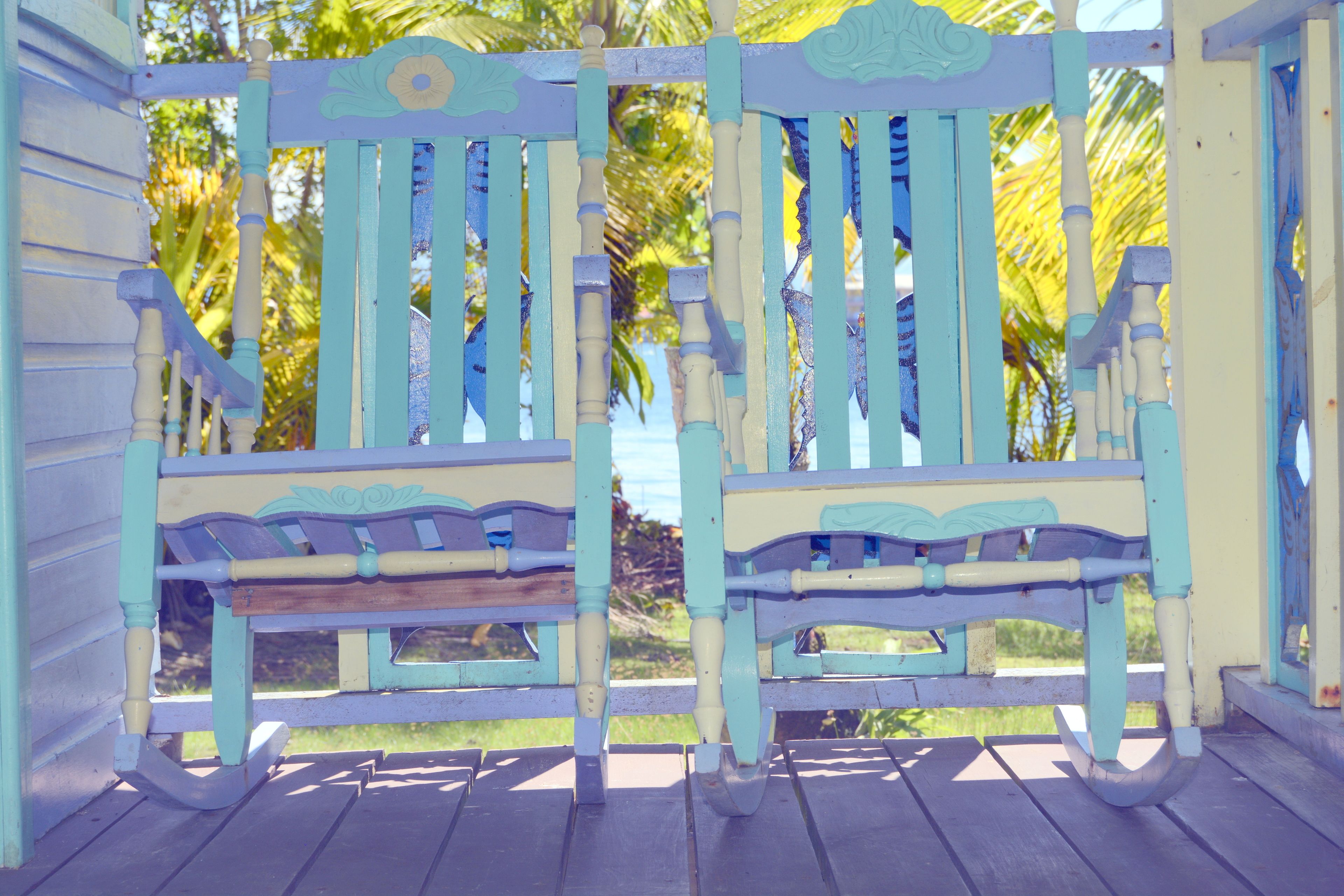 A wooden bench with a light blue and white paint scheme, positioned on a wooden deck with a view of palm trees and a body of water.