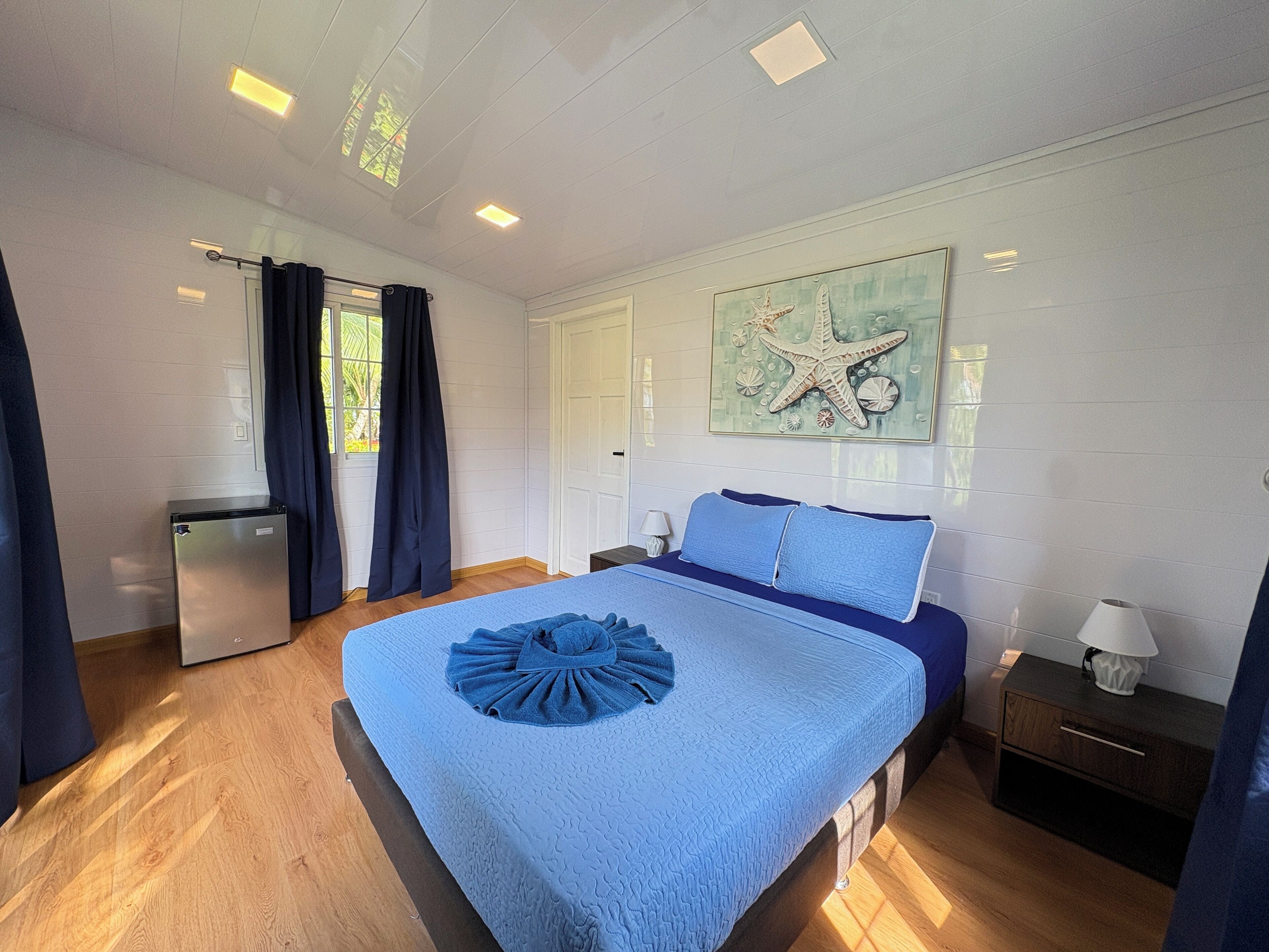 A bedroom with a bed, a small bedside table, a nightstand, a window with curtains, and a wall-mounted artwork.